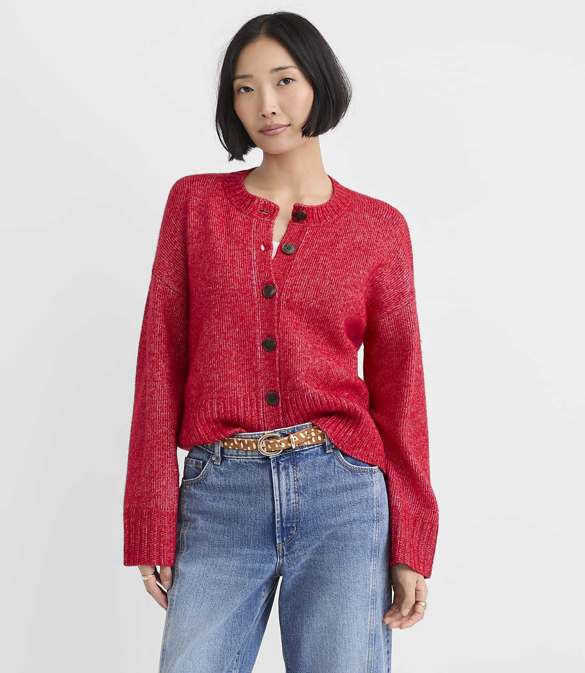 A woman wearing a red button-down cardigan and blue jeans stands against a plain white background.