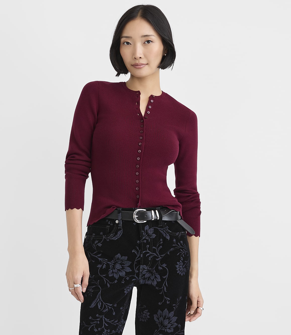 A woman wearing a burgundy button-down top and floral black pants stands against a plain white background.