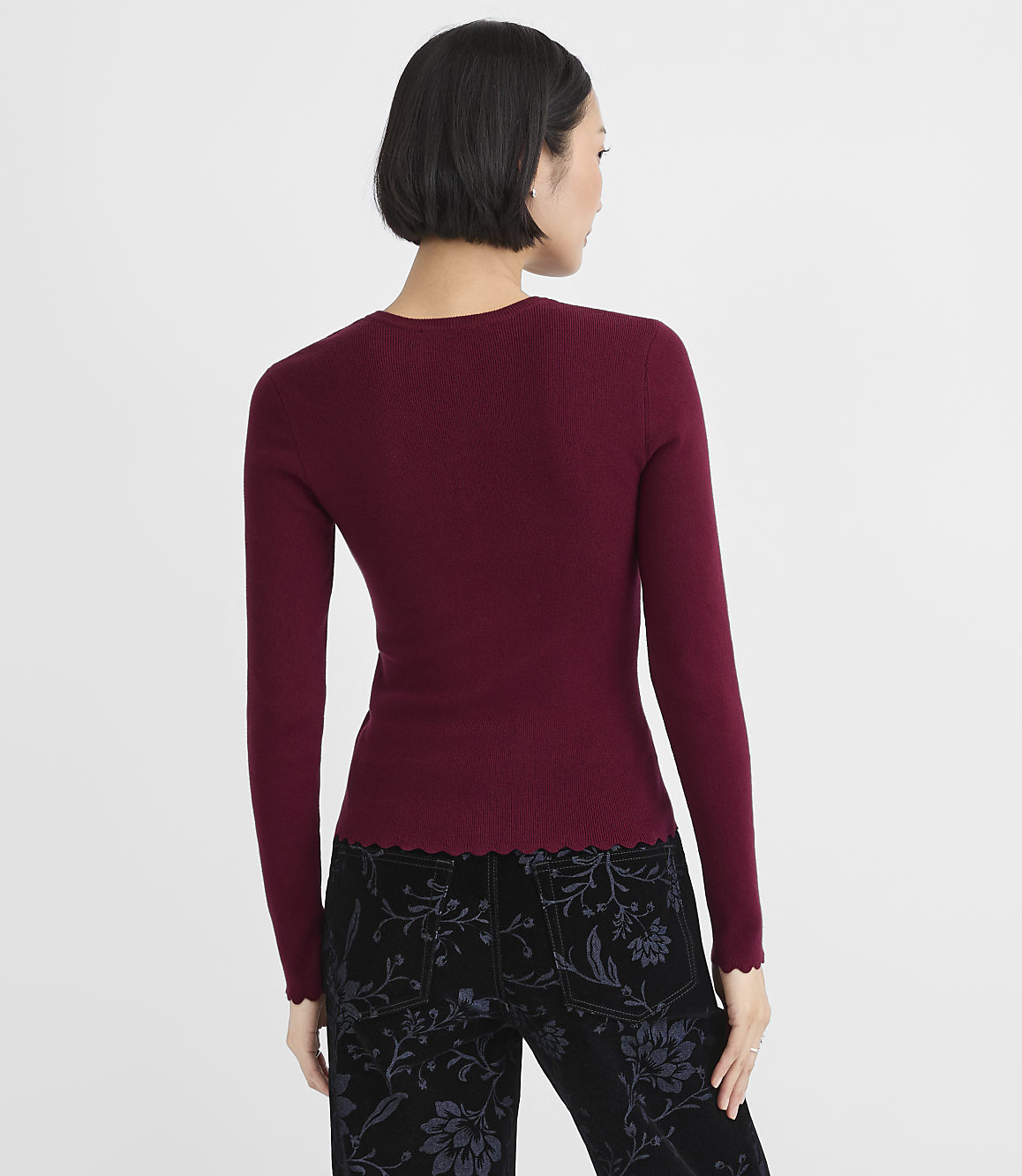 A woman wearing a burgundy long-sleeved top and floral black pants stands with her back to the camera against a plain white background.