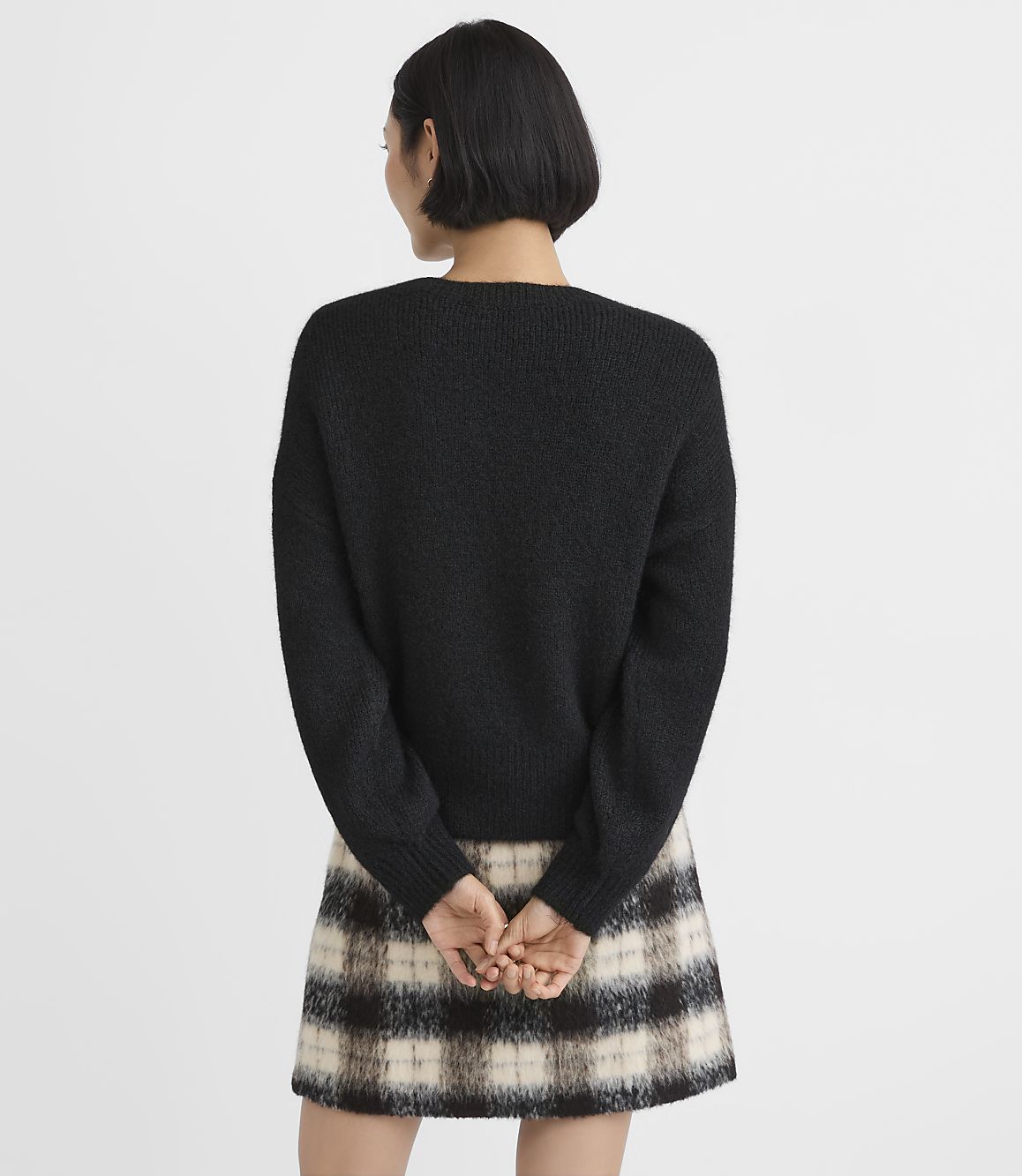 A woman wearing a black sweater and a plaid skirt stands with her back to the camera against a plain white background.