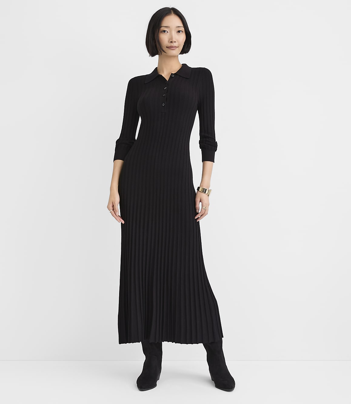 A woman wearing a long-sleeved, pleated black dress and black boots stands against a plain white background.