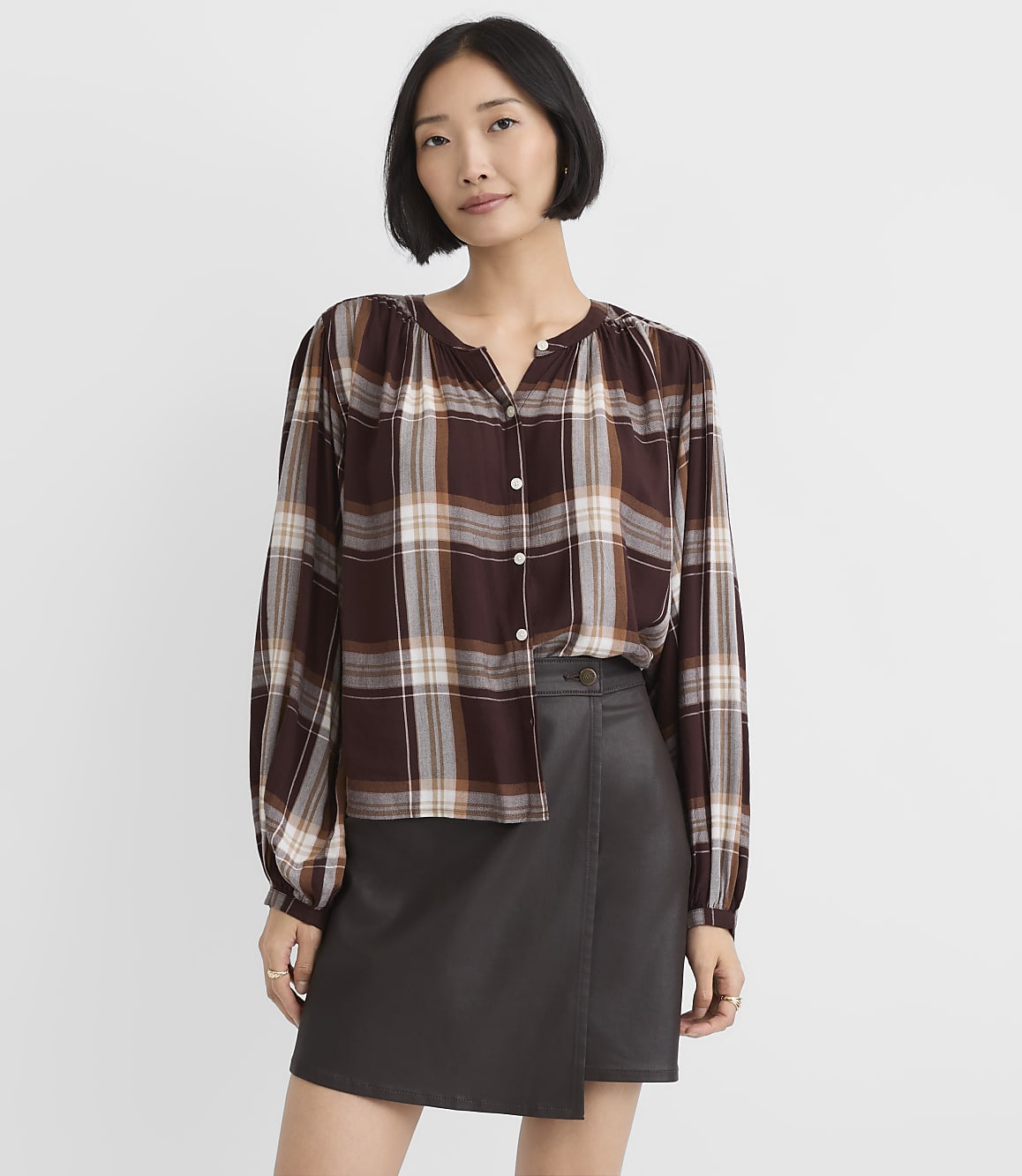 A woman wearing a brown plaid blouse and a dark grey faux-leather skirt stands against a plain white background.