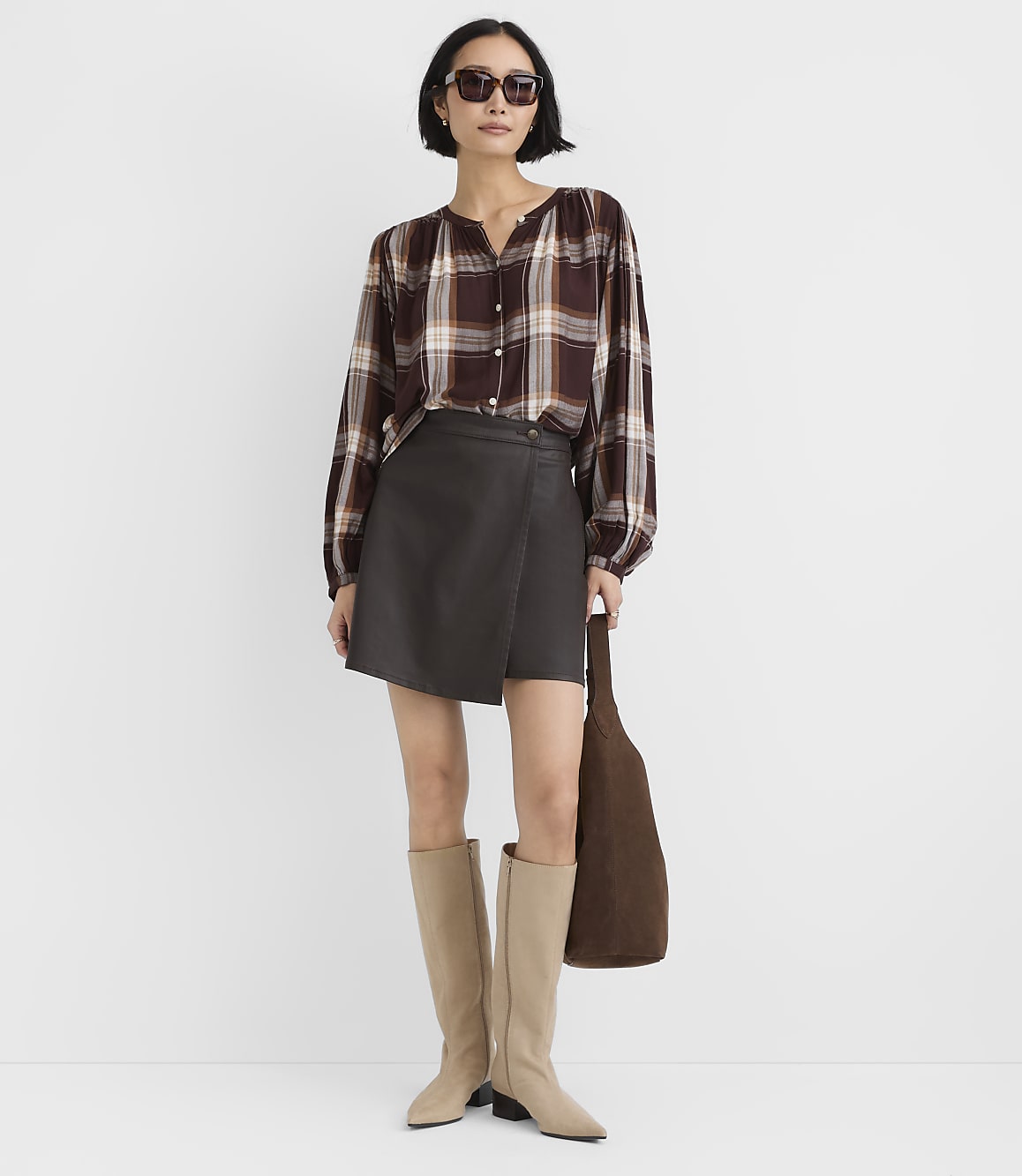 A woman wearing a plaid shirt, a dark grey skirt, and beige boots stands against a plain white background.