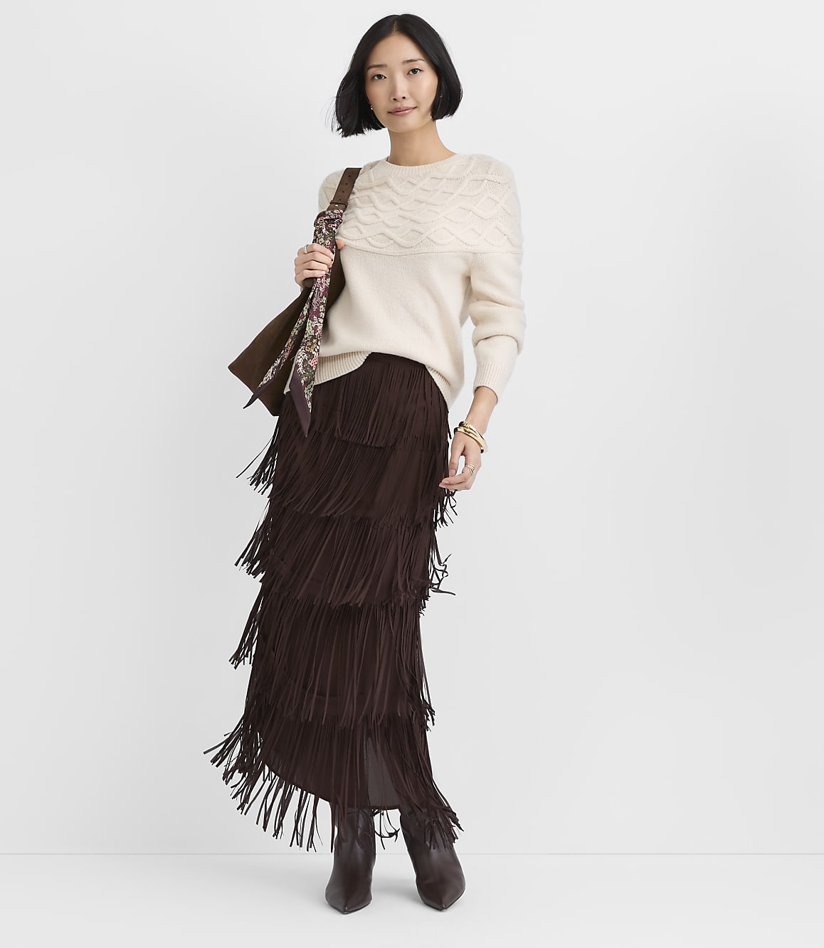 A woman wearing a cream cable-knit sweater and a tiered brown fringe skirt stands against a plain white background.