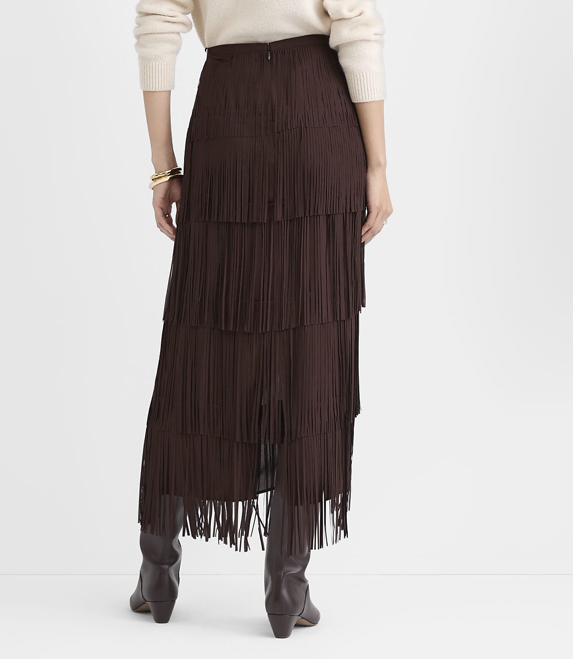 A person wearing a cream sweater and a long, tiered brown fringe skirt with brown boots stands against a plain white background.