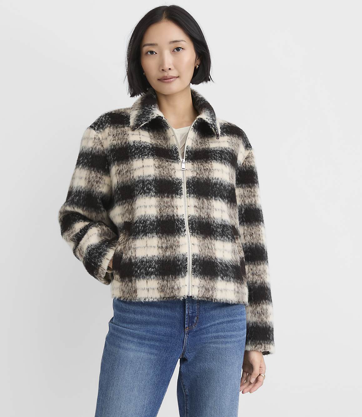 A woman wearing a black and white plaid jacket and blue jeans stands against a plain white background.