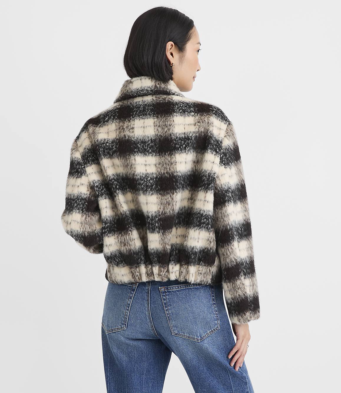 A woman wearing a brown and white checkered faux-fur jacket and blue jeans stands against a plain white background.
