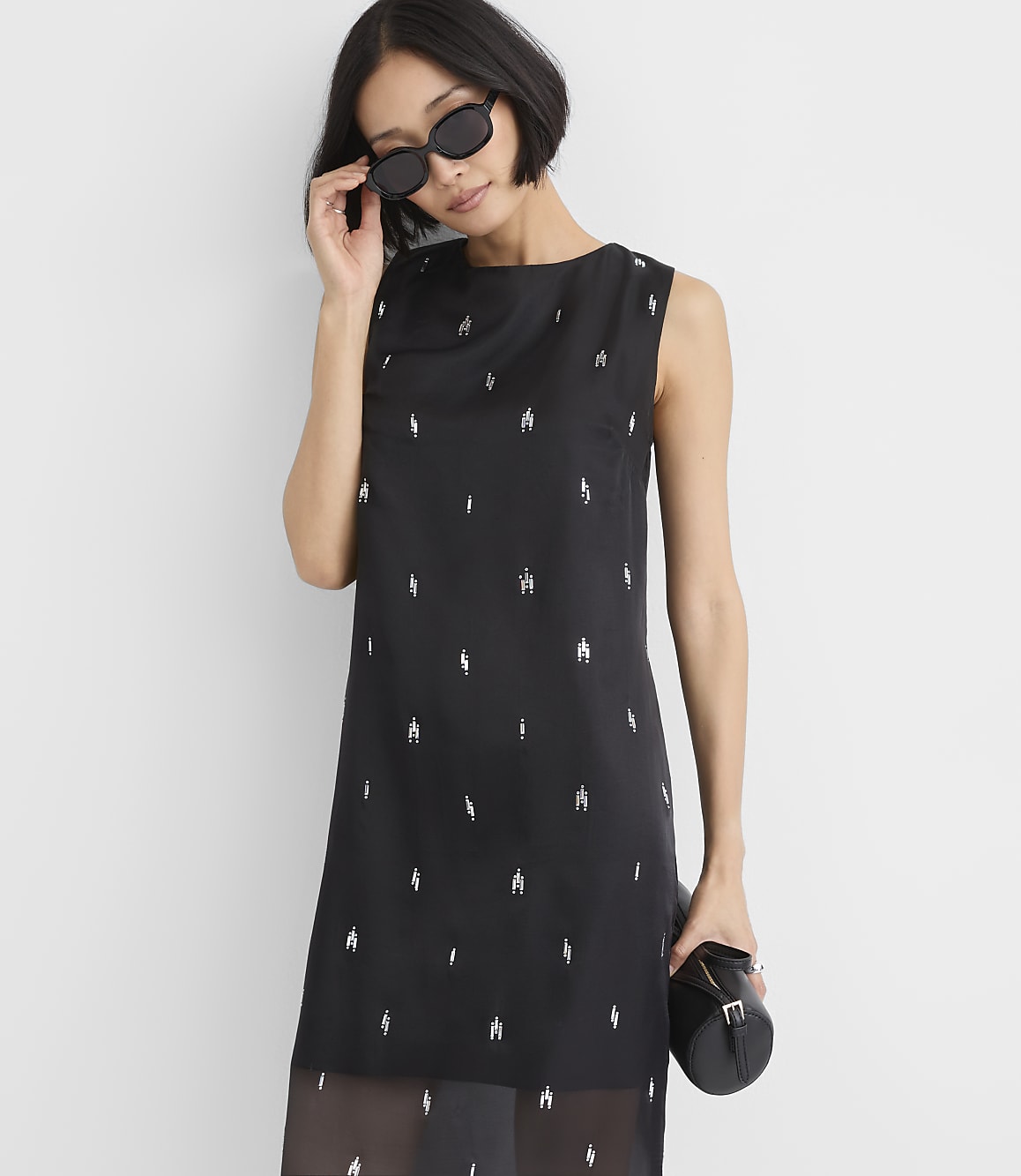 A woman wearing sunglasses and a black sleeveless dress with silver embellishments stands against a plain white background.