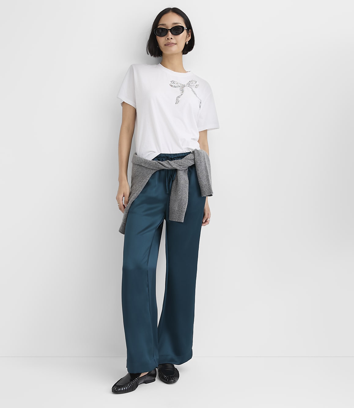 A woman wearing a white t-shirt, teal satin trousers, and sunglasses stands against a plain white background.