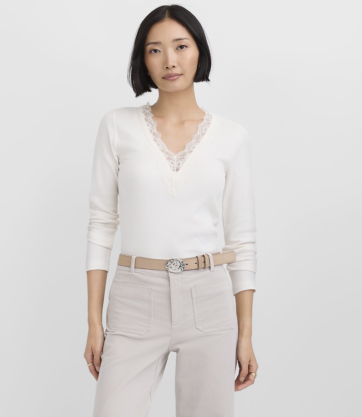 A woman wearing a white lace-trim top and light-colored trousers stands against a plain white background.