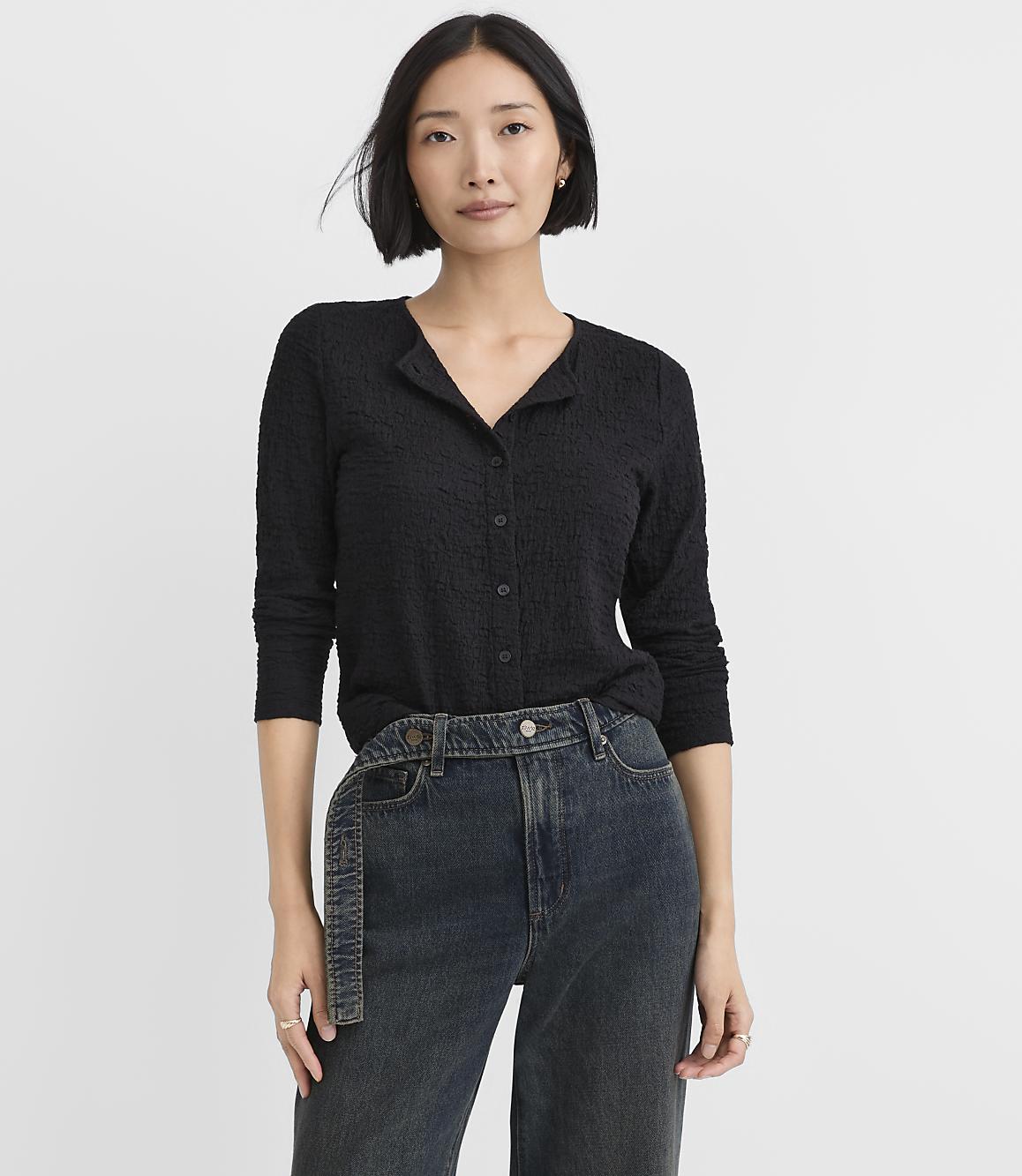 A woman wearing a black textured button-down shirt and dark blue jeans stands against a plain white background.