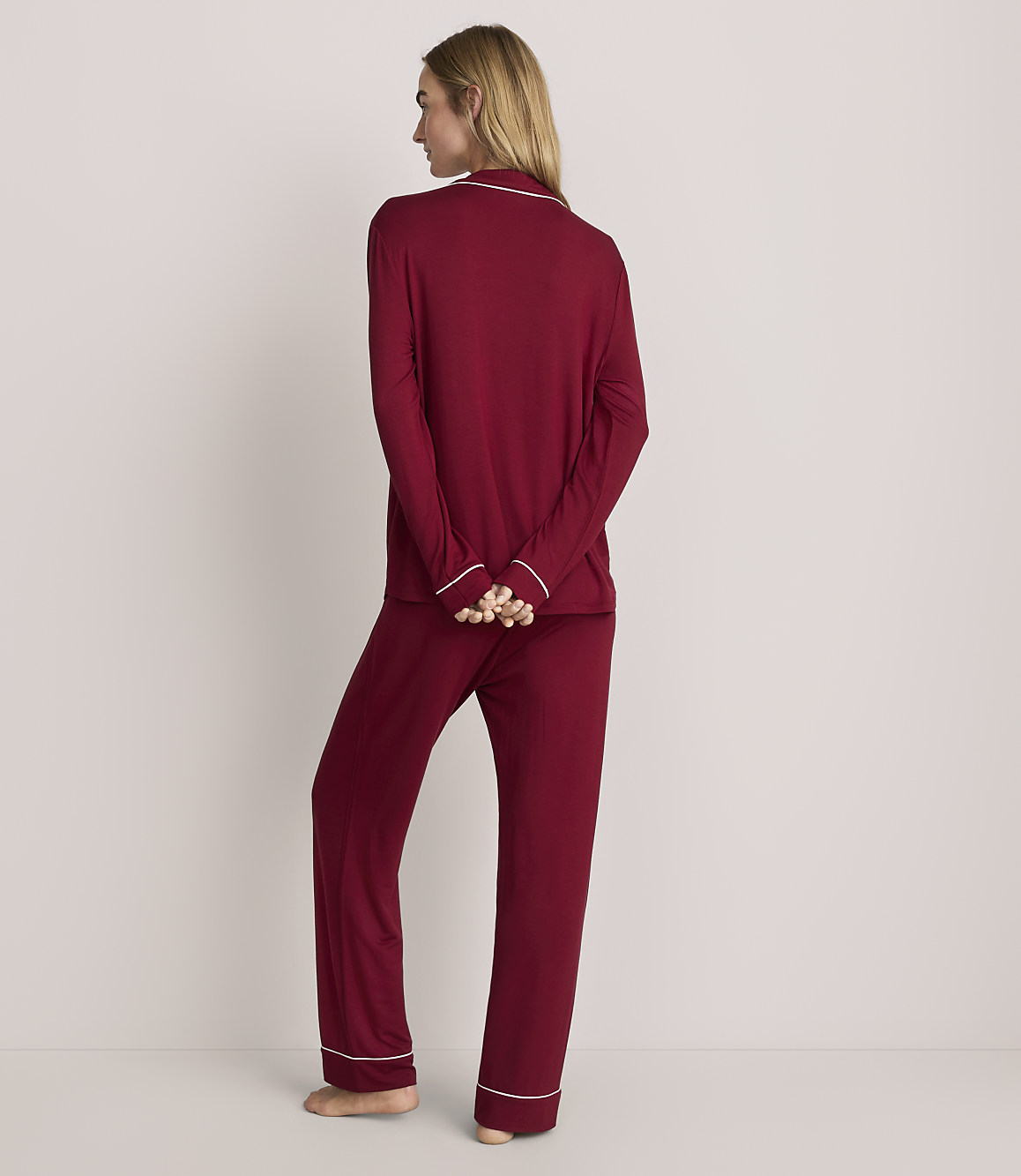 A woman stands with her back to the camera wearing burgundy pajamas with white trim against a plain, light-colored background.