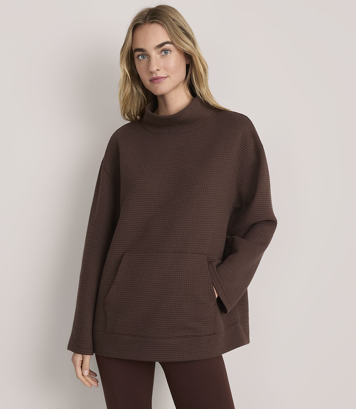 A blonde woman wearing a brown textured oversized tunic and leggings stands against a plain light-colored background.