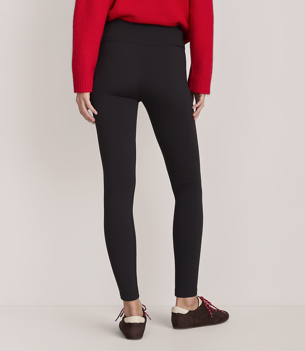 A person wearing a red sweater, black leggings, and brown shoes stands against a plain, light-colored background.