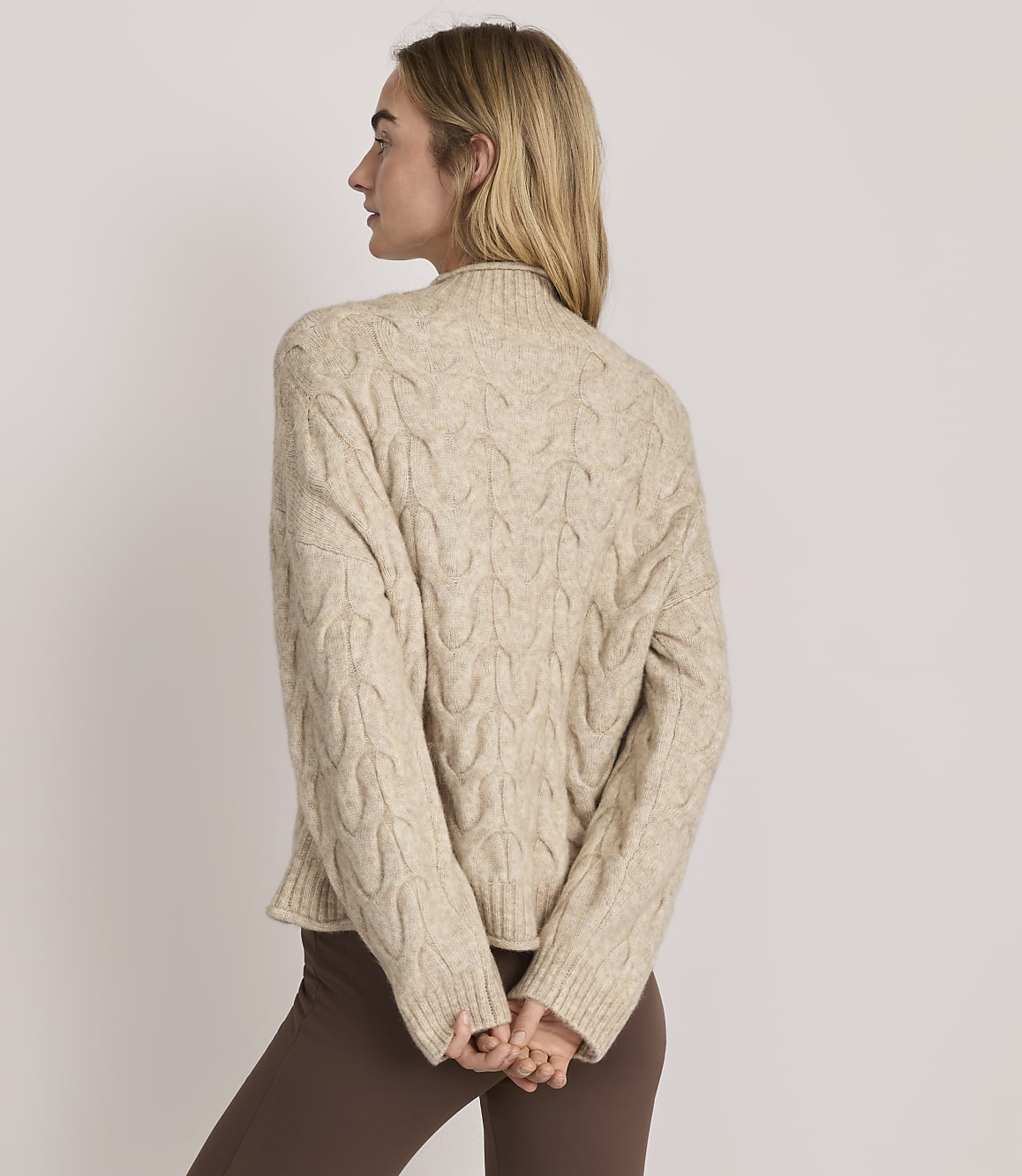 A woman wearing a beige cable-knit sweater and brown pants stands with her back to the camera against a plain light-colored background.