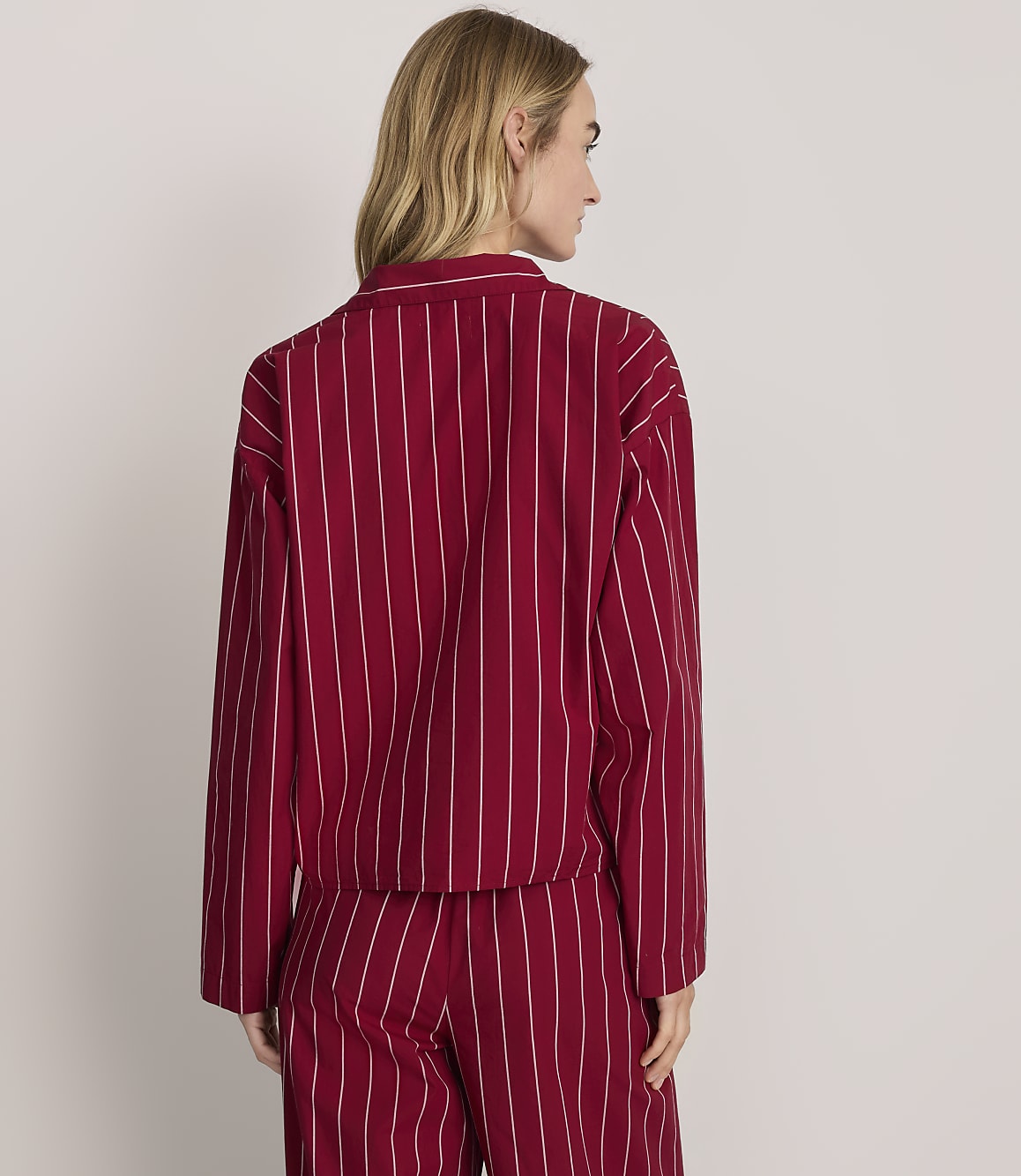 A woman wearing a red and white pinstriped pajama set stands with her back to the camera against a plain light gray background.