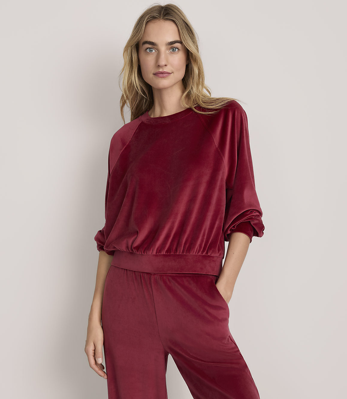 A woman wearing a red velvet two-piece lounge set stands against a plain, light-colored background.