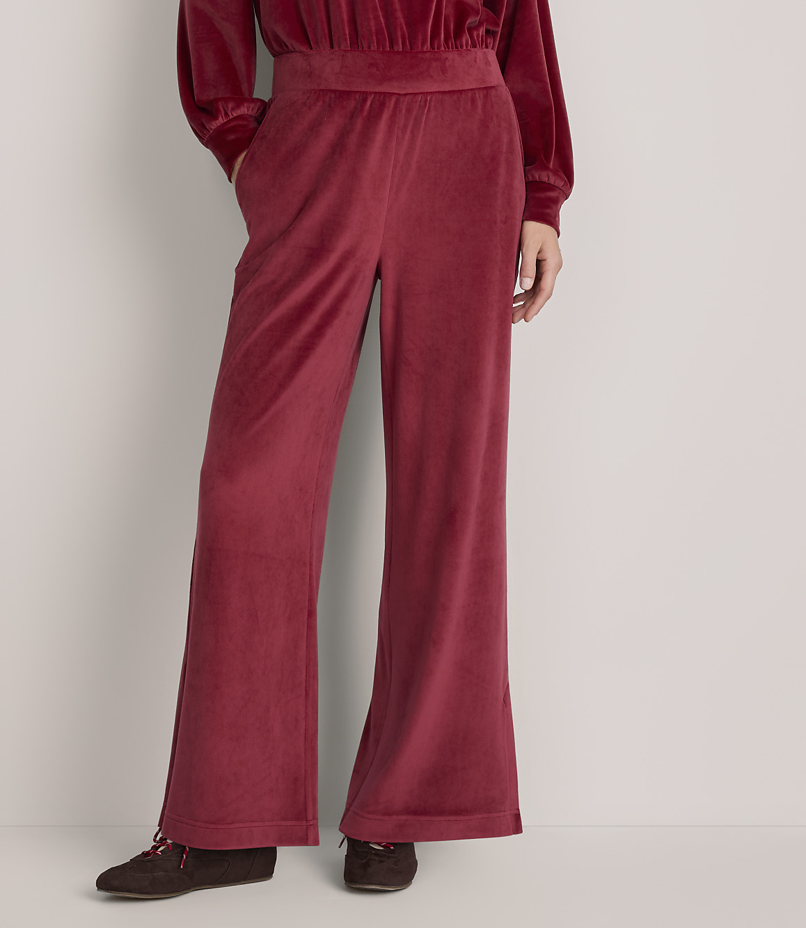 A person wears wide-leg red velvet trousers and a matching top against a plain, light-colored wall.
