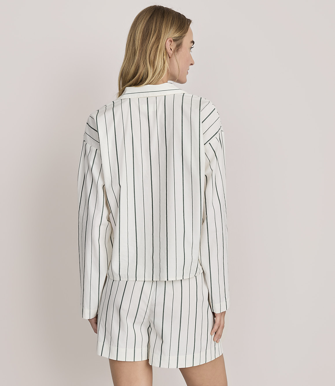 A woman stands with her back to the camera wearing a white and green striped matching shirt and shorts set against a plain light grey background.