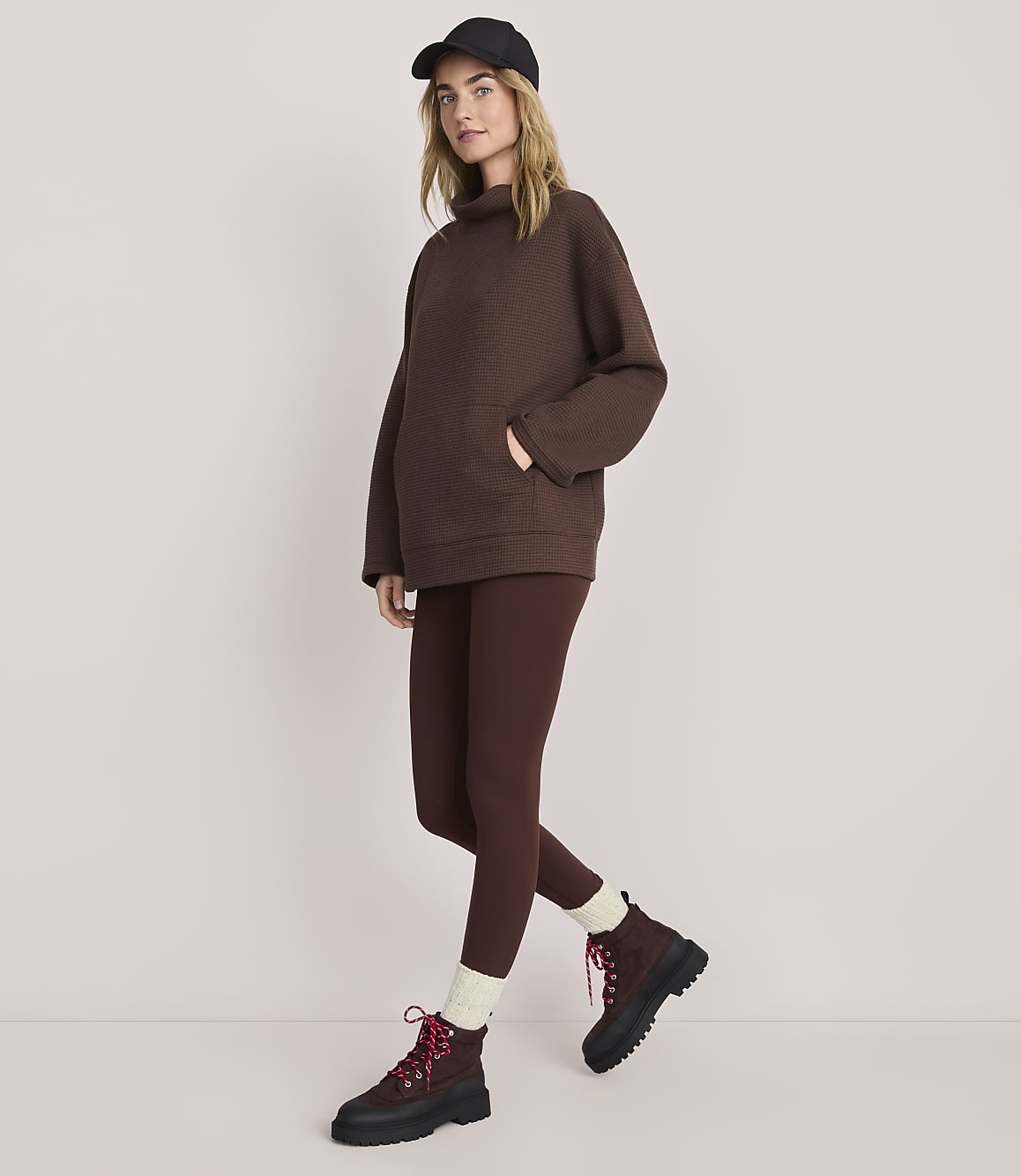 A woman wearing a brown oversized sweater, matching leggings, a black cap, and brown boots stands against a plain, light-colored background.