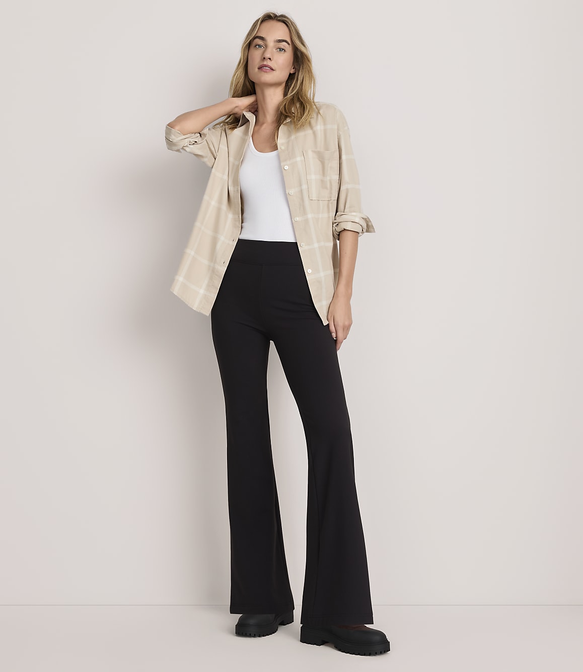 A woman wearing a white top, a beige plaid shirt, and black flared trousers stands against a plain, light-colored background.