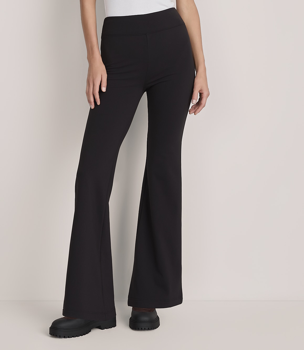 A person wearing black flared trousers and chunky boots stands against a plain, light-colored background.