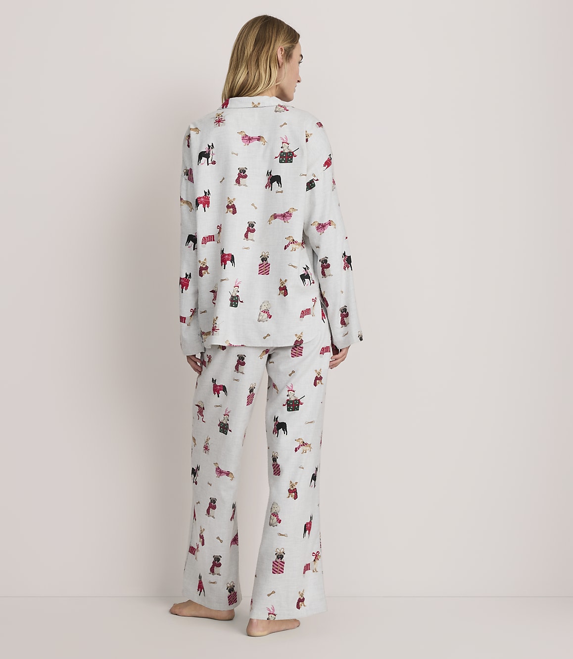 A woman stands with her back to the camera wearing light grey pajamas with a dog print against a plain, light-colored background.