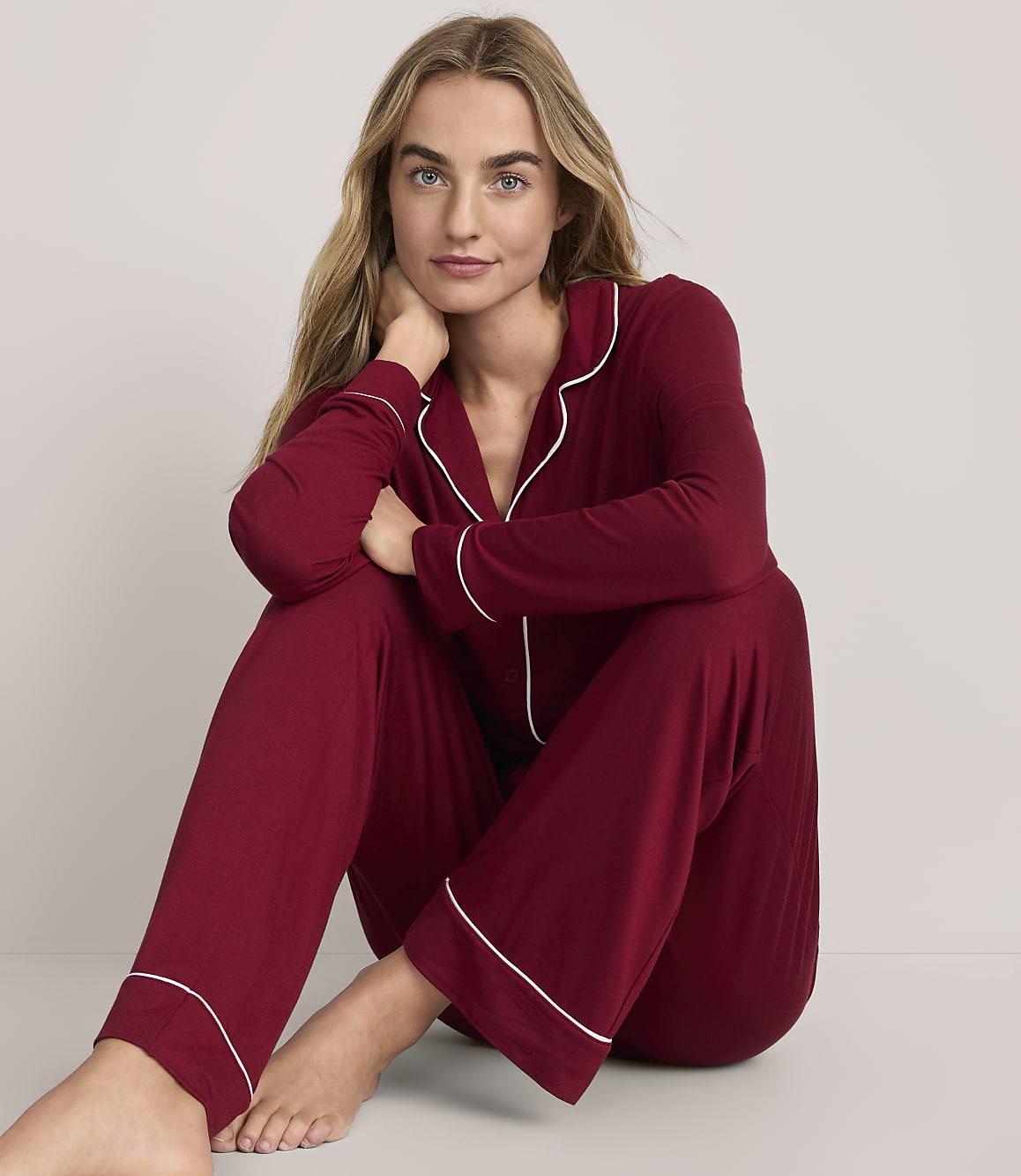 A woman wearing burgundy pajamas with white piping sits on the floor against a plain, light-colored background.