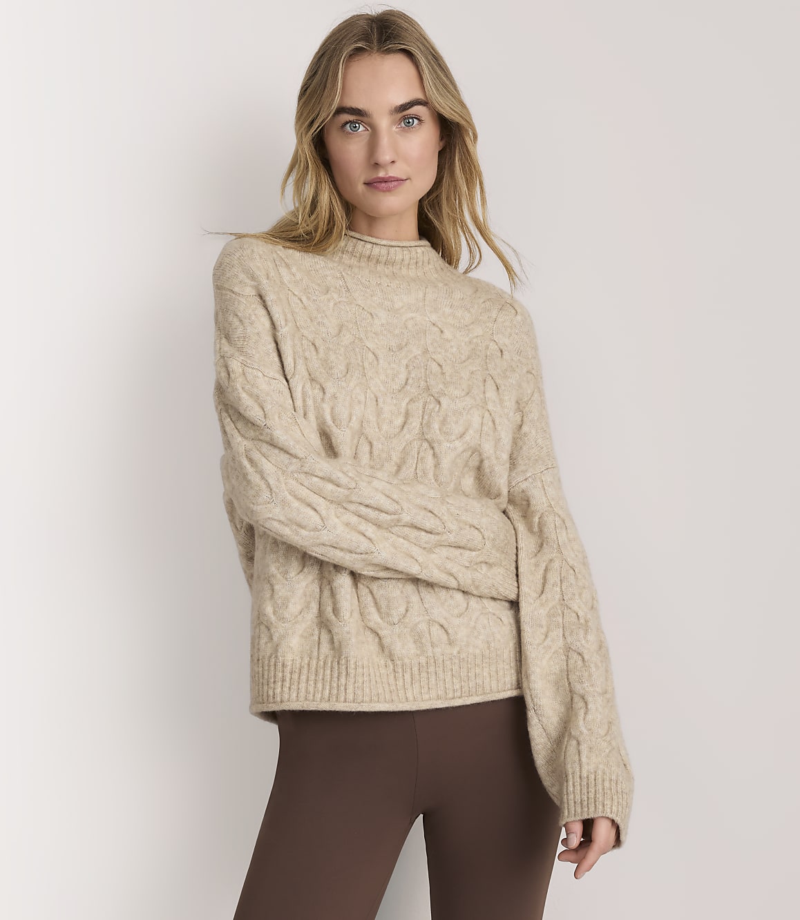 A woman wearing a beige cable-knit sweater and brown leggings stands against a plain white background.