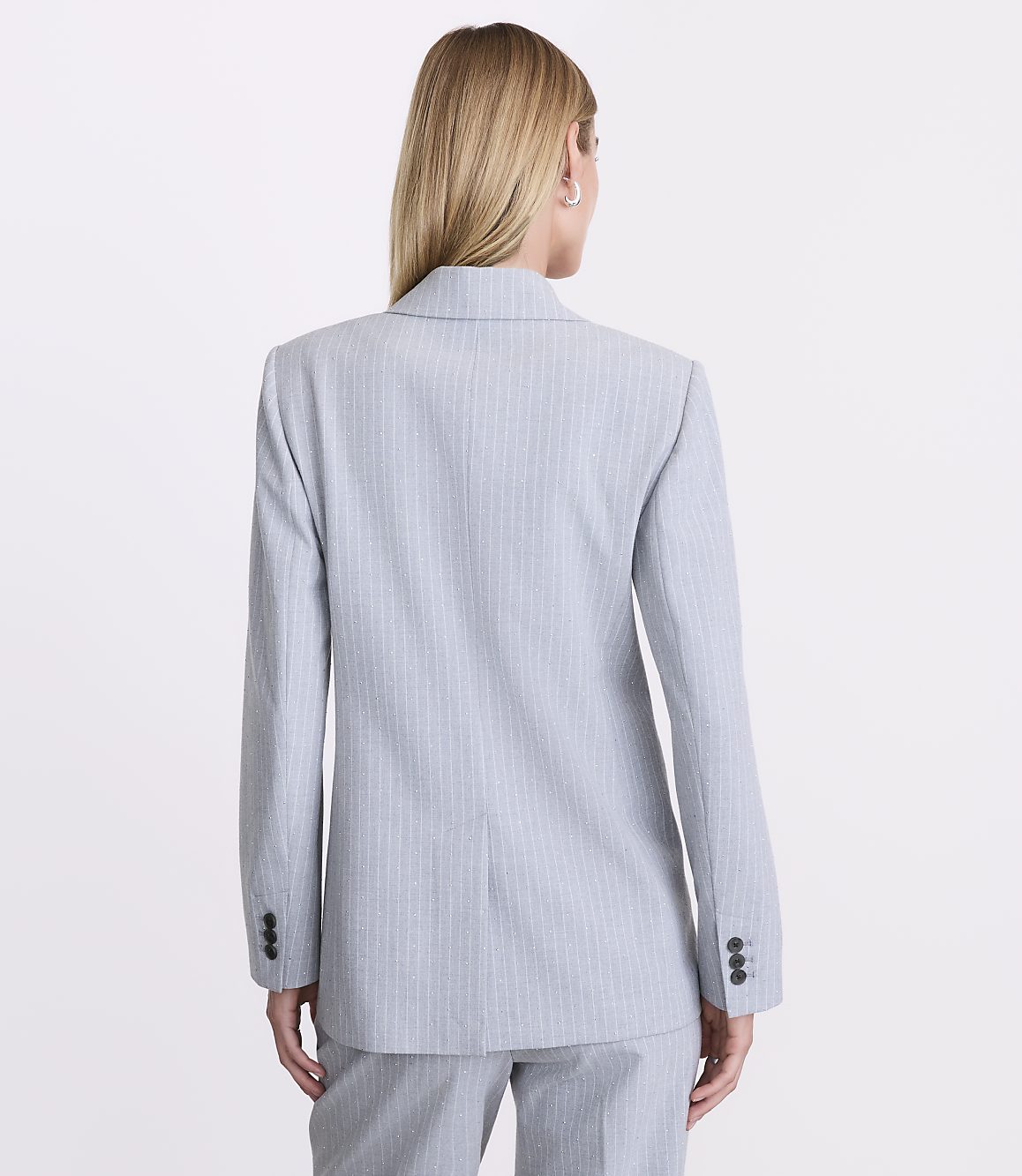 A woman with blonde hair stands with her back to the camera wearing a light blue pinstriped blazer and matching trousers against a plain white background.