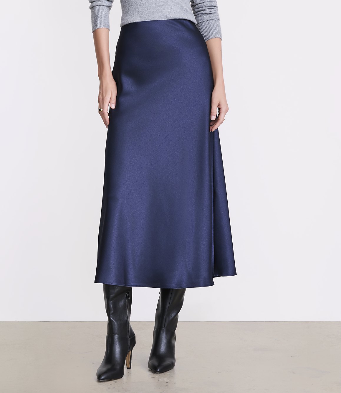 A person wears a long navy blue satin skirt, a grey sweater, and black leather boots against a plain white background.
