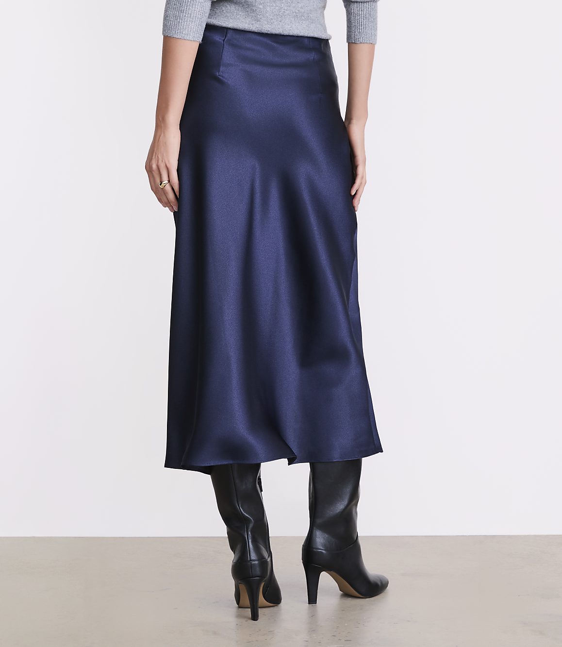 A person wearing a grey top, a long navy blue satin skirt, and black heeled boots stands against a plain white background.
