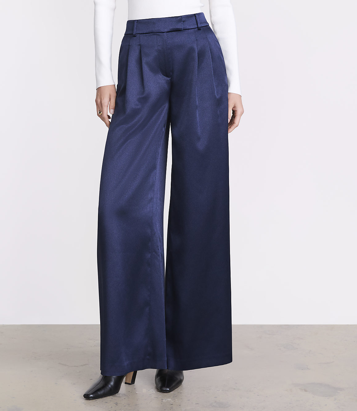 A person wears wide-leg navy blue trousers, a white long-sleeved top, and black pointed heels against a plain white background.