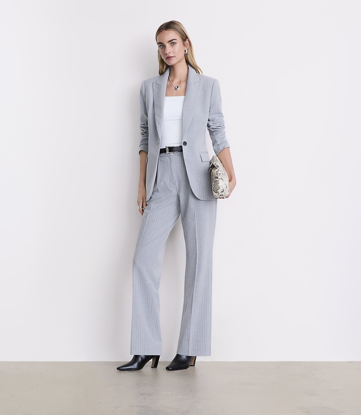 A woman wearing a light grey pinstripe suit and black heels stands against a plain white background.