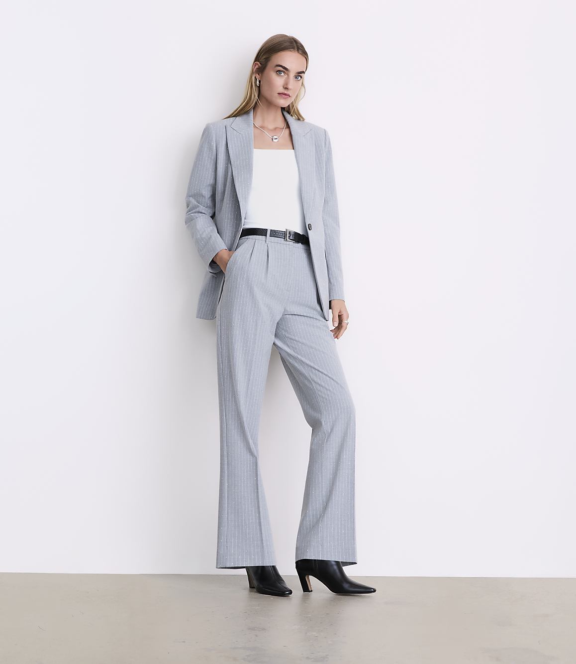 A woman wearing a light blue pinstripe suit and black heels stands against a plain white wall.