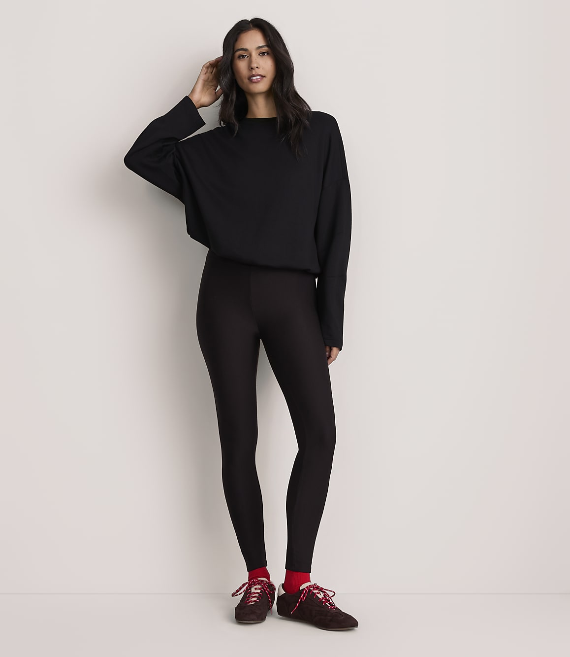 A woman wearing a black long-sleeved top, black leggings, and maroon sneakers with red socks stands against a plain white background.