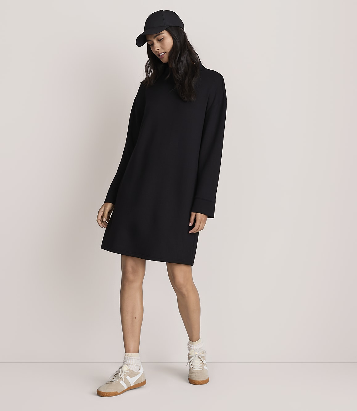 A woman wearing a black baseball cap, a long-sleeved black dress, and beige sneakers stands against a plain, light-colored background.