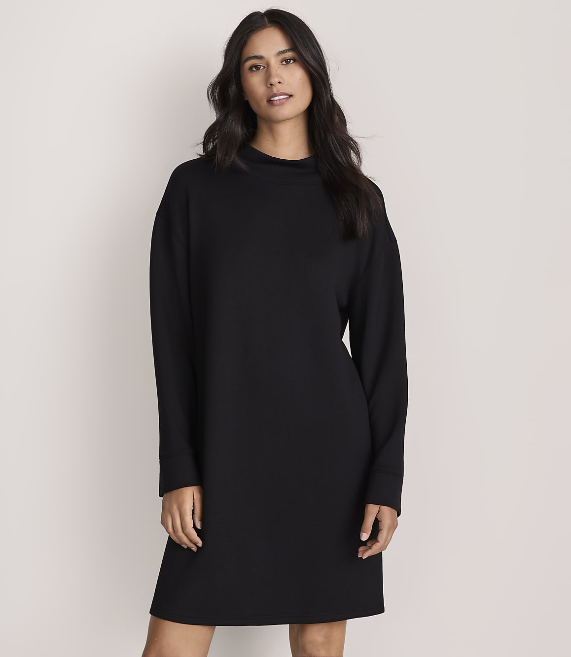 A woman with long dark hair wearing a long-sleeved black dress stands against a plain, light-colored background.