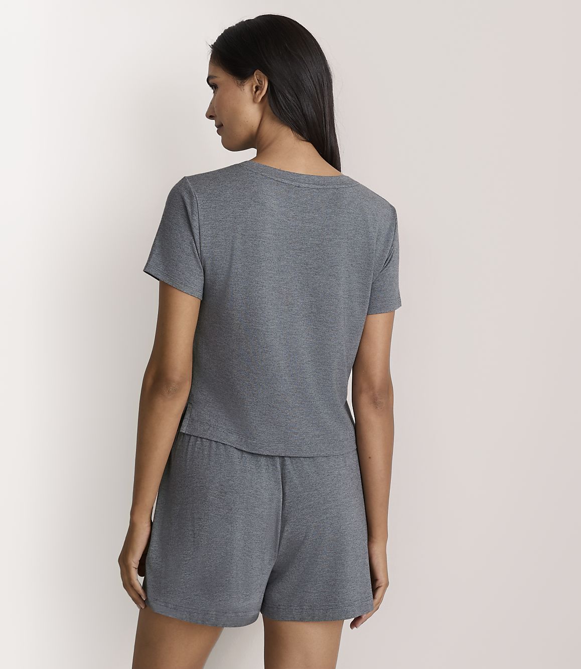 A woman wearing a grey short-sleeved t-shirt and matching shorts stands with her back to the camera against a plain white background.