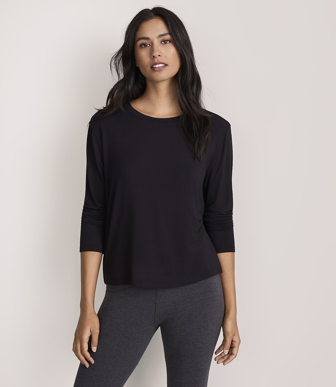 A woman wearing a black long-sleeved top and dark grey leggings stands against a plain, light-colored background.