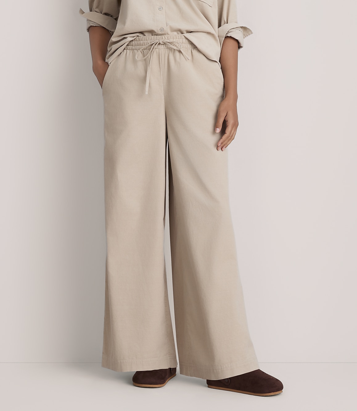 A person wearing a beige button-down shirt and wide-leg beige trousers stands against a plain white background.