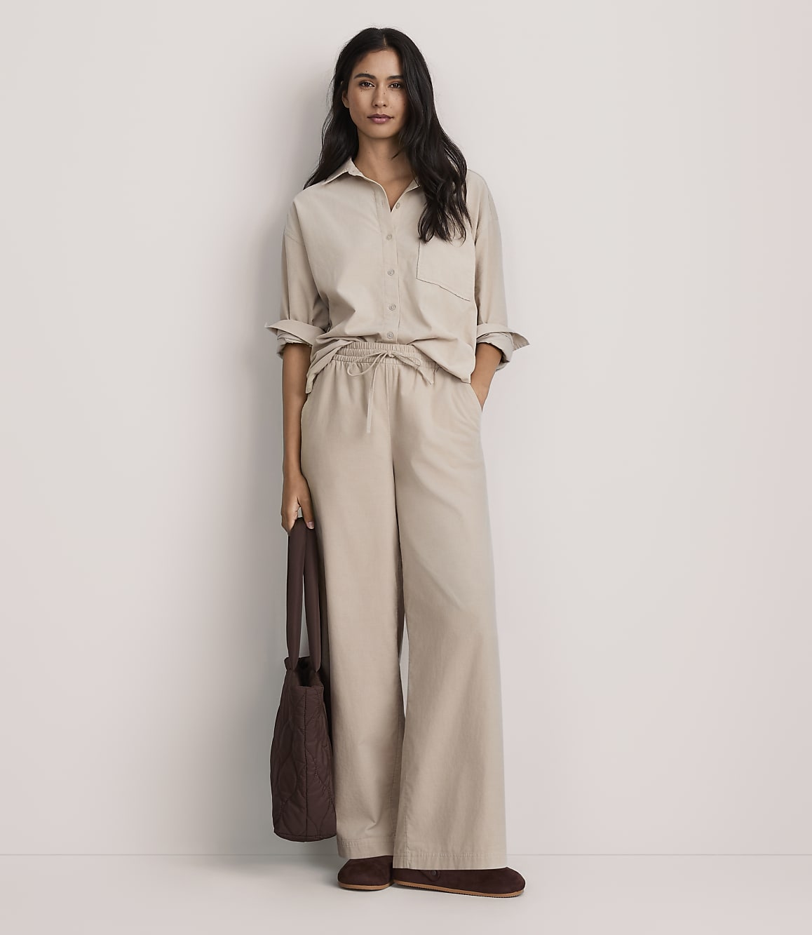 A woman wearing a beige button-down shirt and matching wide-leg trousers stands against a plain white background while holding a brown handbag.