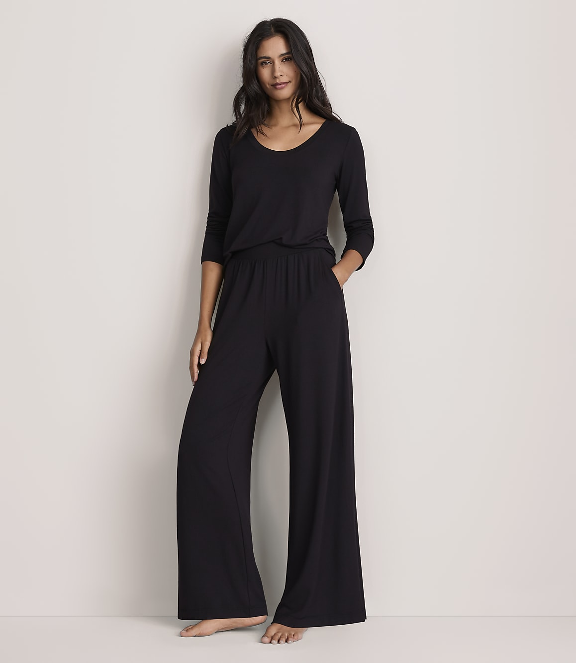 A woman wearing a black long-sleeved top and wide-leg black trousers stands against a plain, light-colored wall.