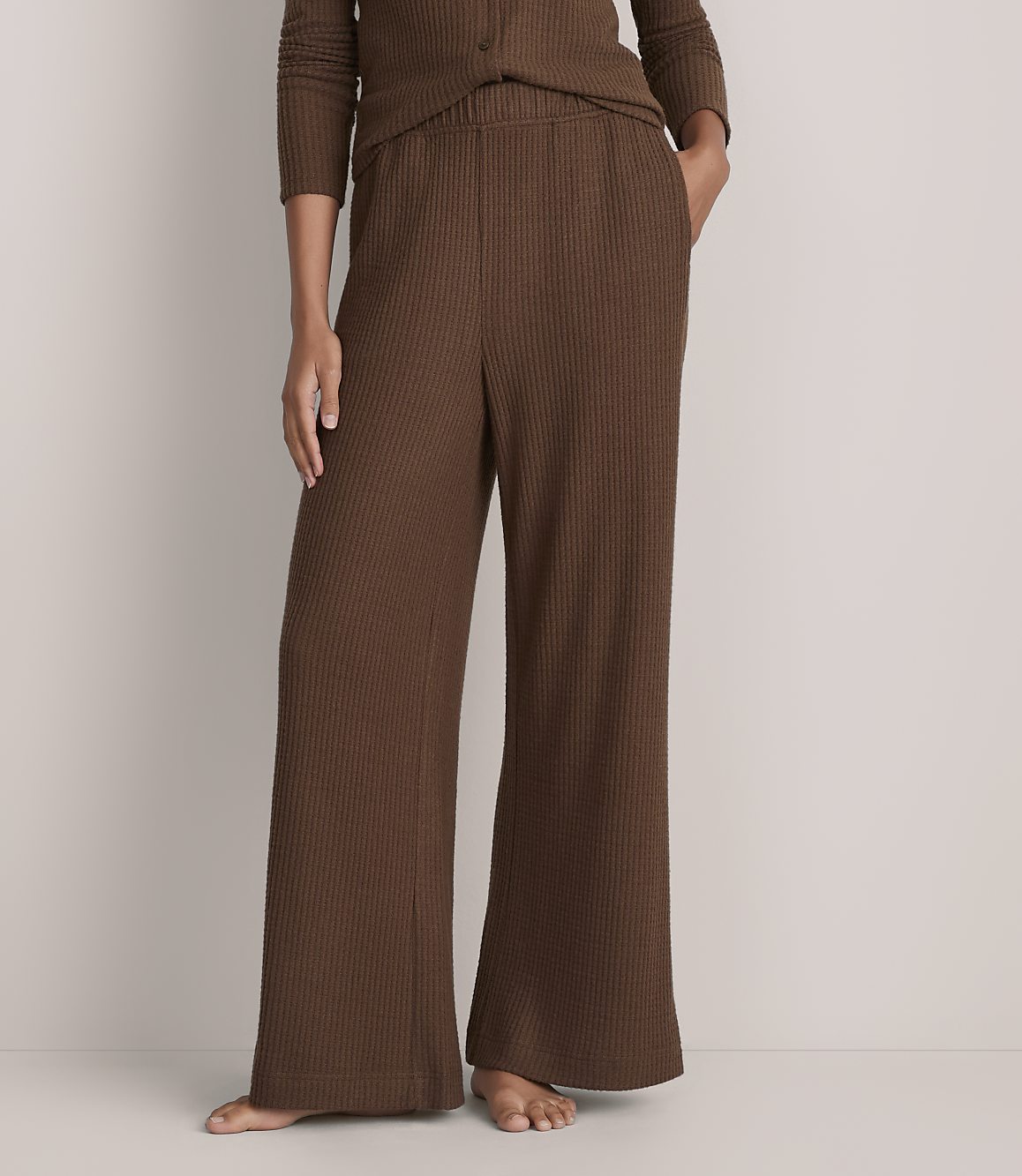 A person wears a brown ribbed knit lounge set against a plain, light-colored wall.