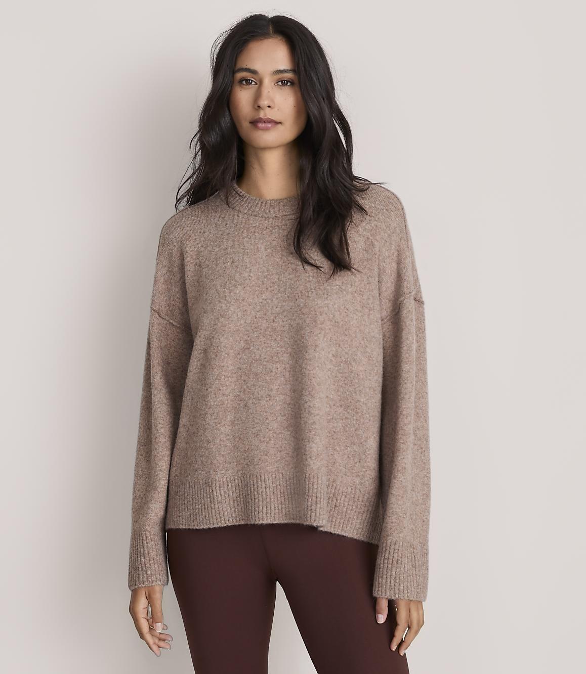 A woman wearing a brown oversized sweater and dark brown leggings stands against a plain, light-colored background.