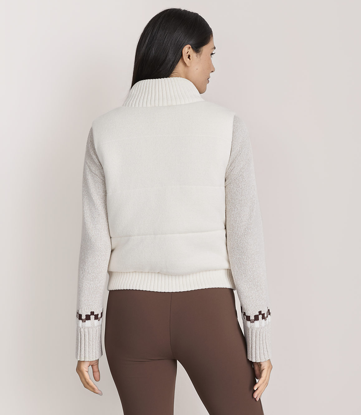 A woman wearing a white textured sweater and brown leggings stands with her back to the camera against a plain light-colored background.
