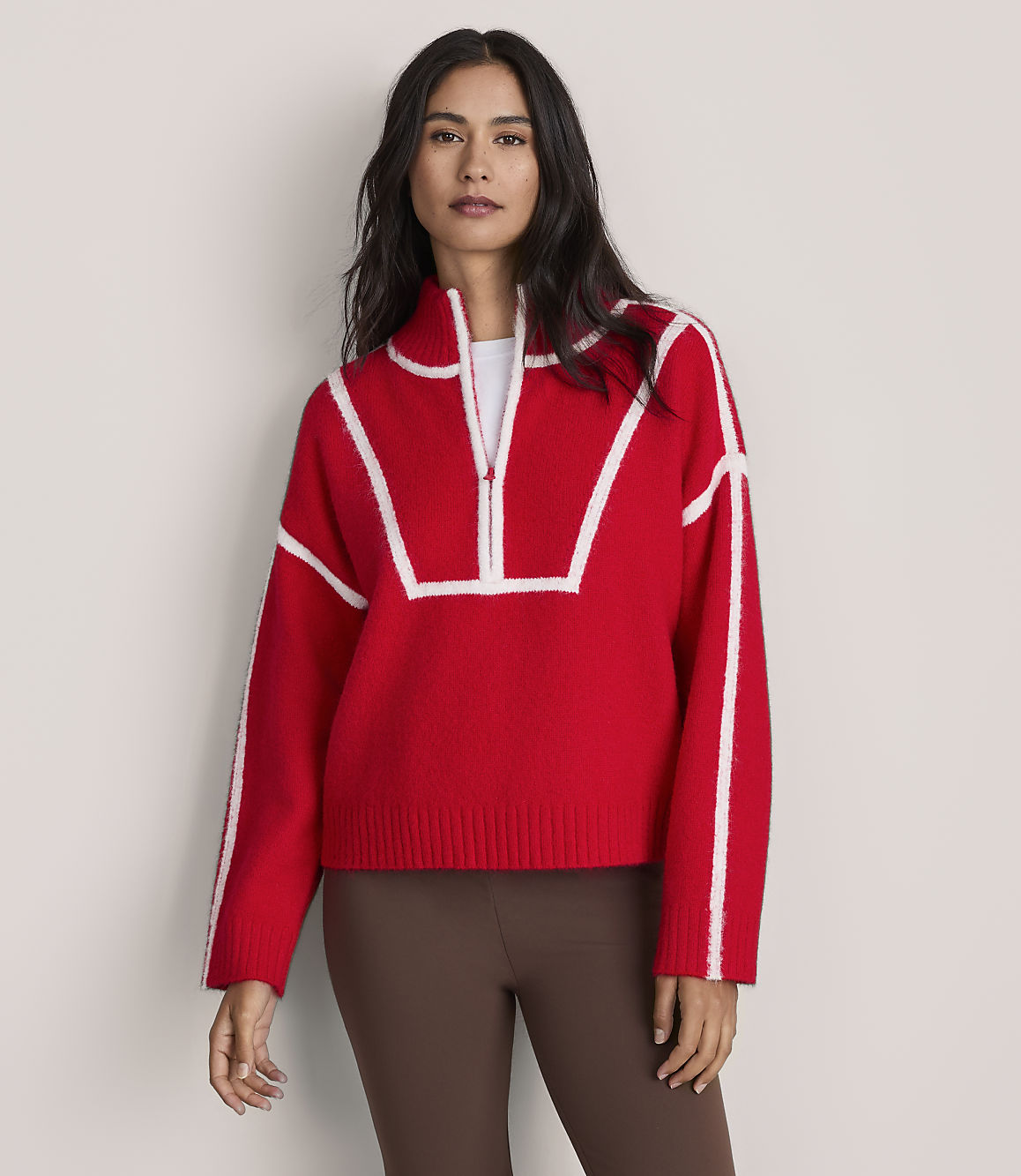 A woman wearing a red and white quarter-zip sweater and brown leggings stands against a plain, light-colored background.