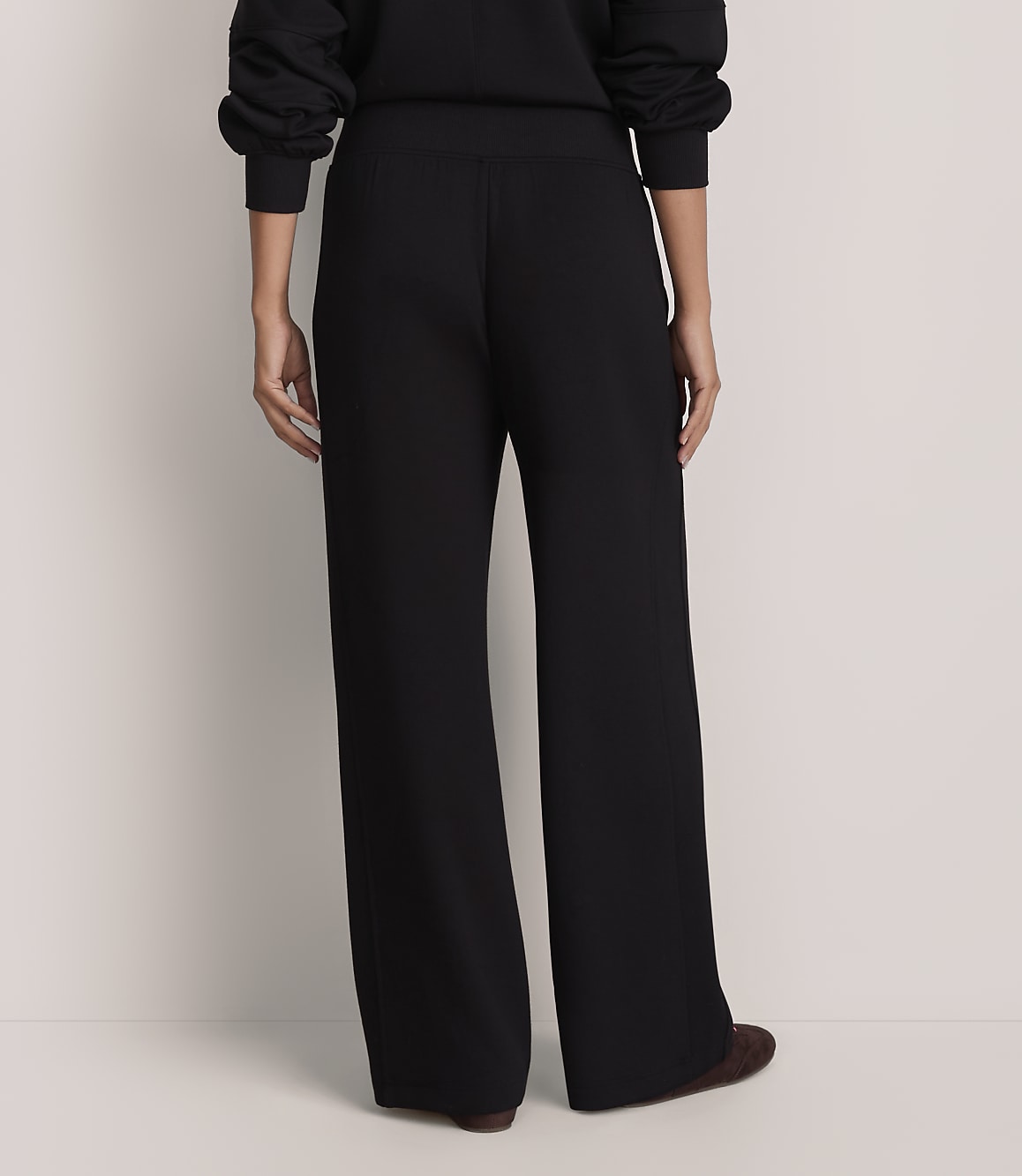 A person stands with their back to the camera wearing black wide-leg trousers and a black top against a plain white background.