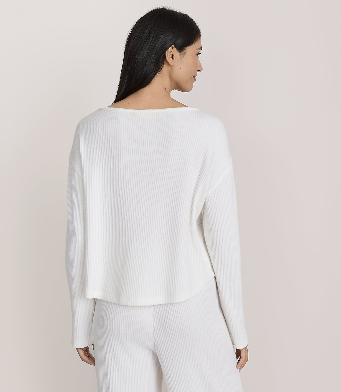 A woman with long dark hair wearing a white ribbed long-sleeved top and matching pants stands against a plain light-colored background.