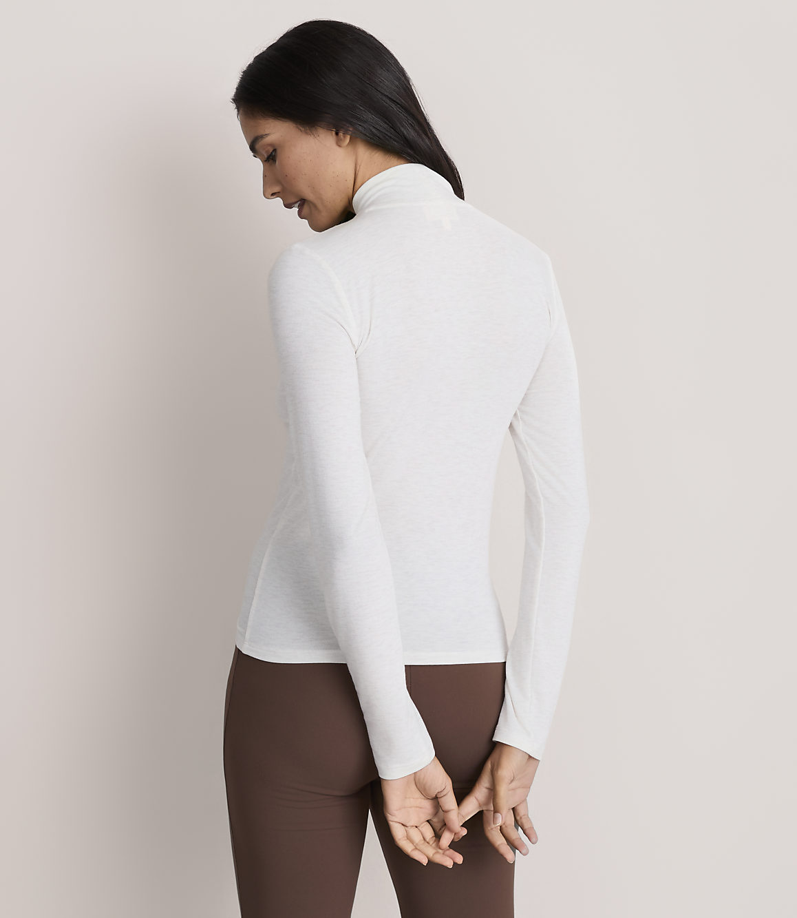 A woman wearing a white long-sleeved turtleneck and brown leggings stands against a plain, light-colored background.